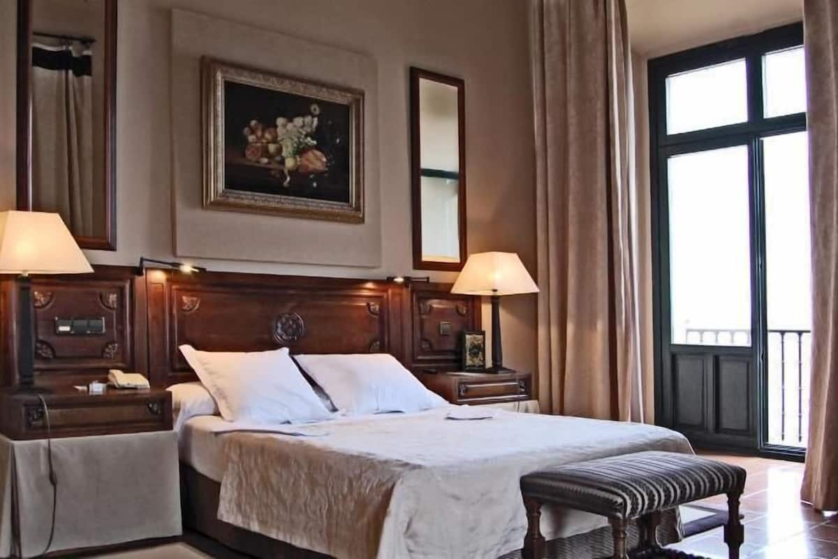 Hotelkamer met een groot bed, twee nachtkastjes, een bankje, een schilderij, een spiegel en een raam met gordijnen.