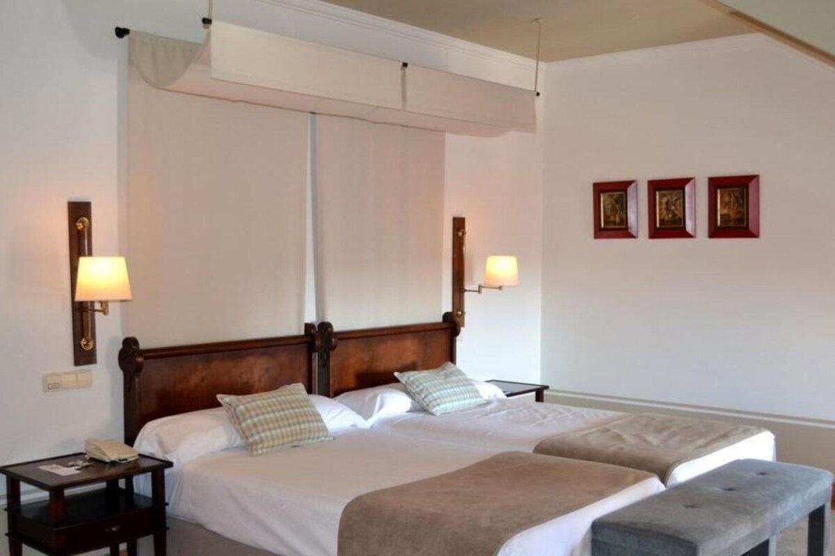 A double bed with a wooden headboard, two bedside tables with lamps, a bench, and three framed pictures on the wall.