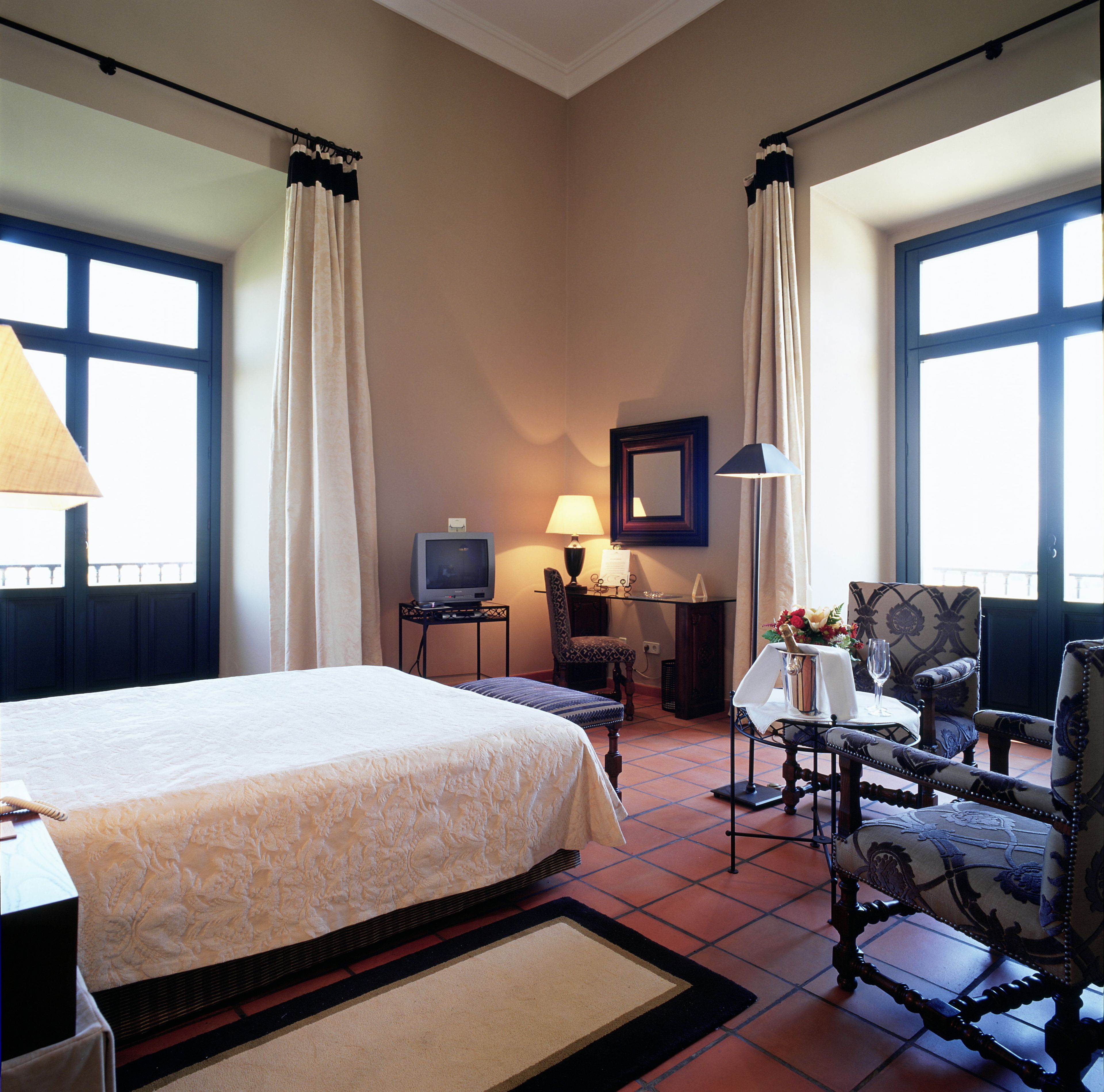 A hotel room with a large bed, a desk with a television, a chair, and a table with a vase of flowers.