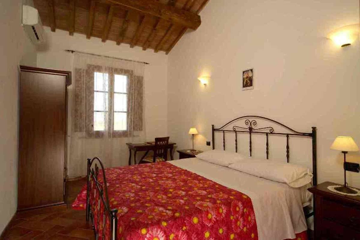 A bedroom with a wooden ceiling, a bed with a floral bedspread, two bedside lamps, a nightstand, a desk, and a window with curtains.