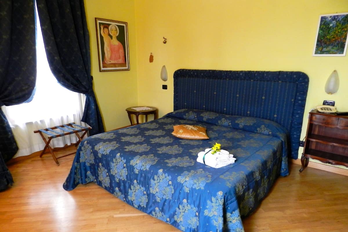 A bedroom with a bed, a small table, a chair, a window with curtains, and a painting on the wall.