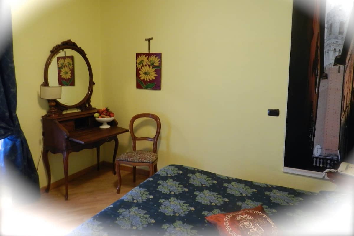 A room with a bed, a wooden chair, a desk with a mirror, a bowl of fruit, and a painting on the wall.