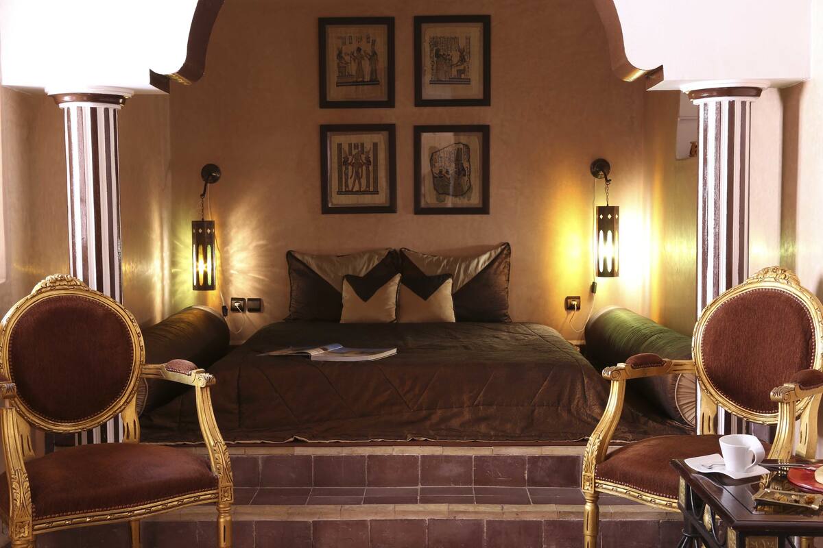 A hotel room with a large bed, two ornate chairs, a small table with a cup and saucer, and four framed pictures on the wall.