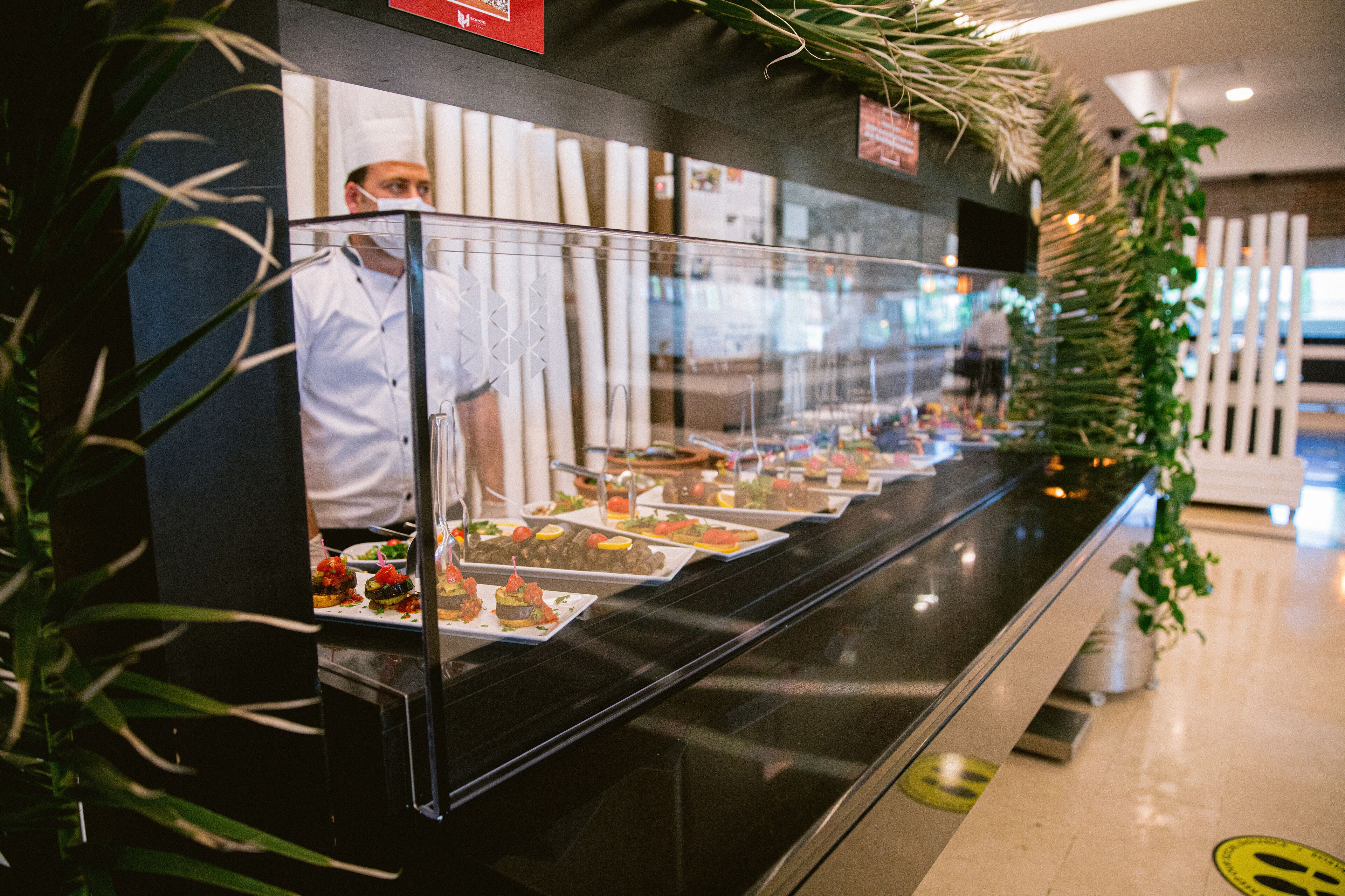 A buffet counter with various dishes displayed in a glass case, surrounded by greenery.