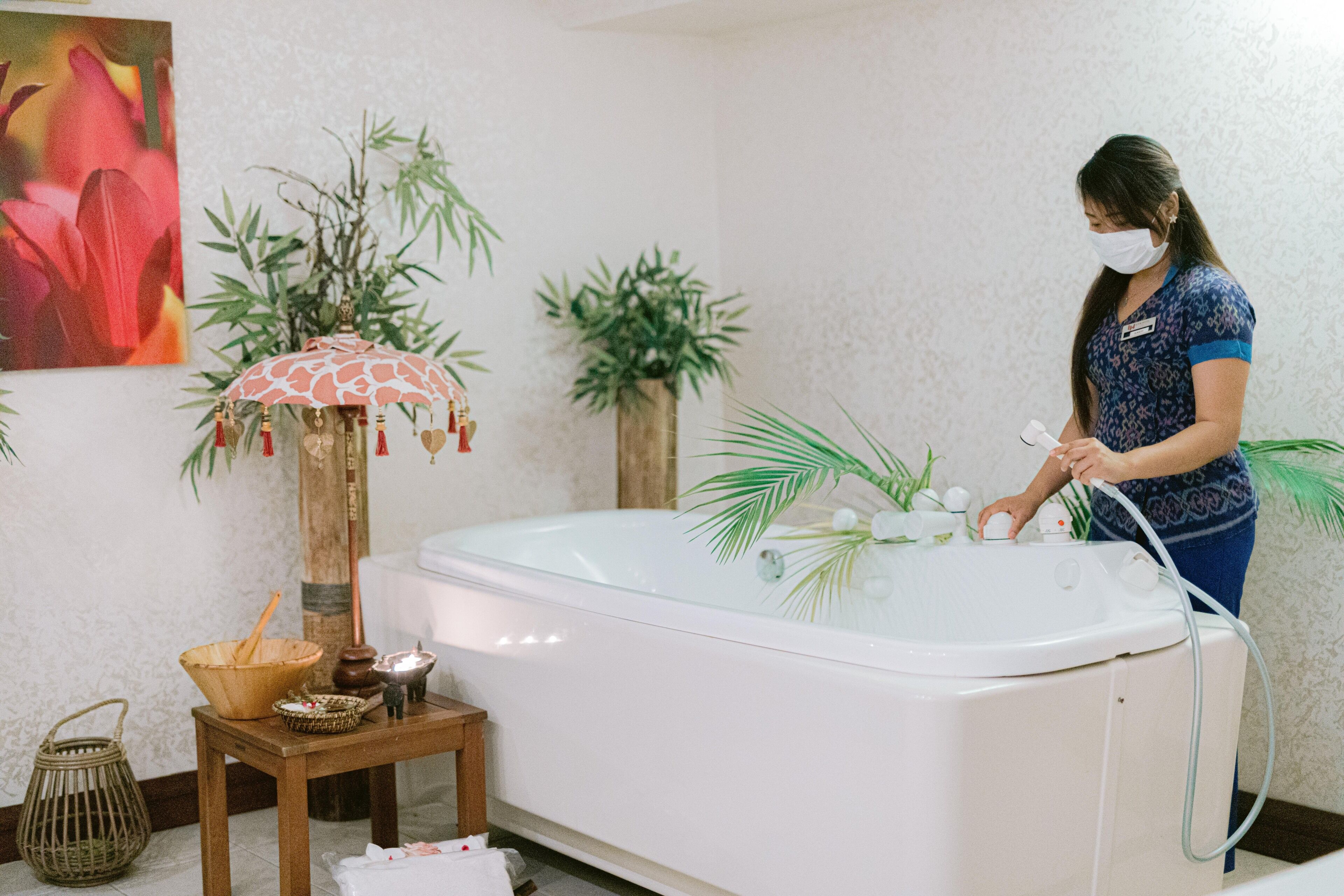 A spa room with a large bathtub, a wooden stool with a bowl, a small table with candles, and a decorative umbrella.