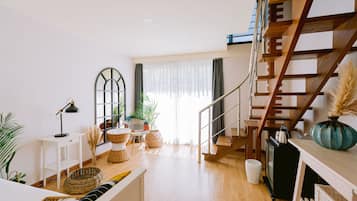 A spacious living area with a wooden staircase, a white sofa, a dining table, and a large window with sheer curtains.