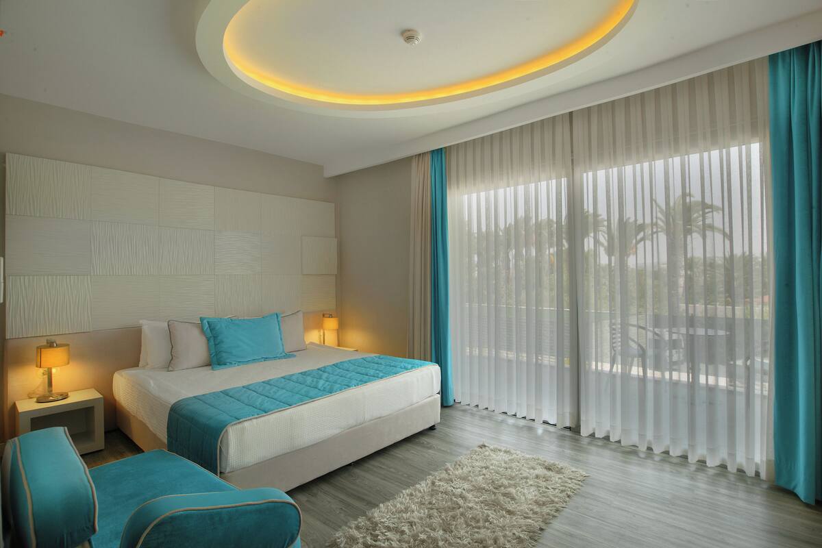 A modern hotel room with a bed, bedside lamps, a view through sheer curtains, and a fluffy rug.