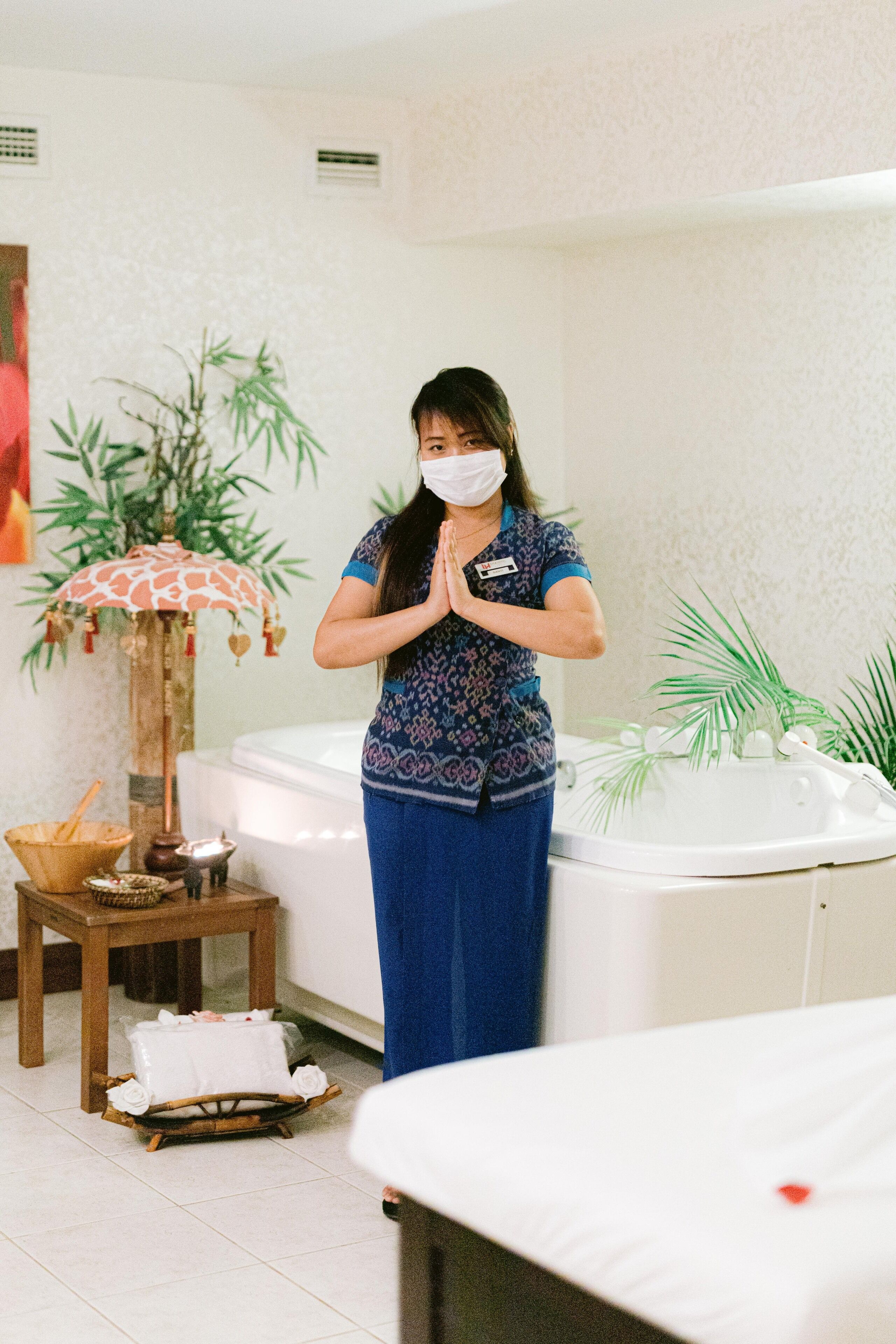Une personne se tient dans une pièce où se trouvent une table de massage, un jacuzzi et une petite table sur laquelle sont posés un panier et une bougie.