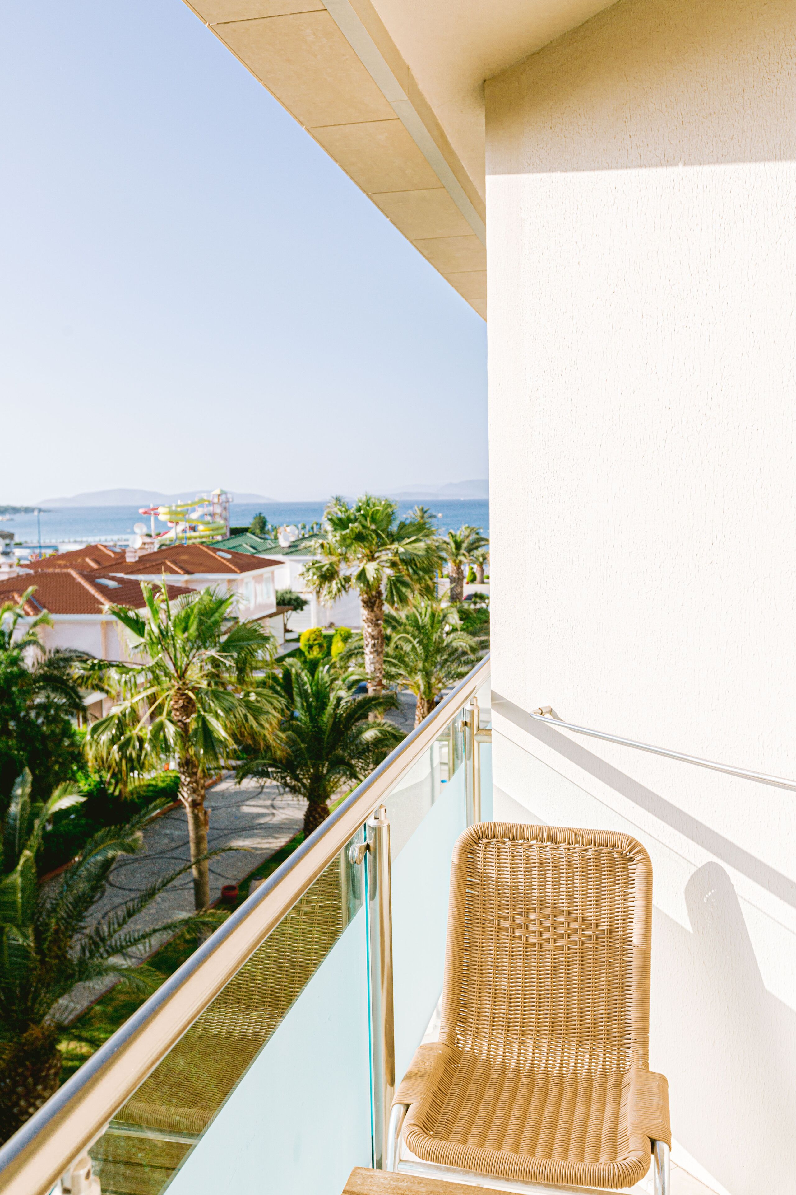 A balcony with a wicker chair, overlooking a coastal area with palm trees and buildings.
