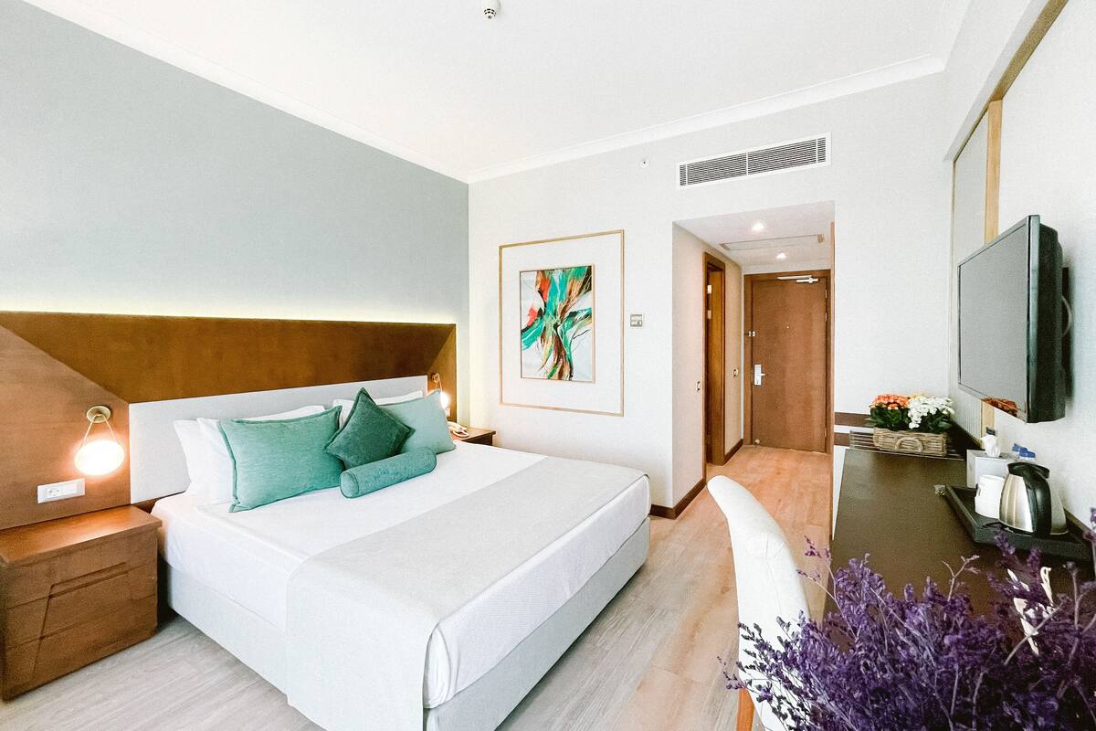 Una habitación de hotel moderna con una cama grande, una mesita de noche con lámpara, un televisor de pantalla plana y un pequeño escritorio con cafetera.