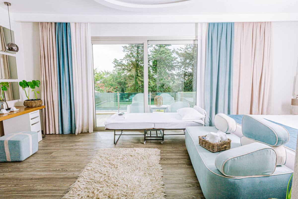 Una habitación de hotel moderna con un amplio ventanal corredero que da a un balcón, un sofá, una mesita pequeña y una mesita de noche con lámpara.