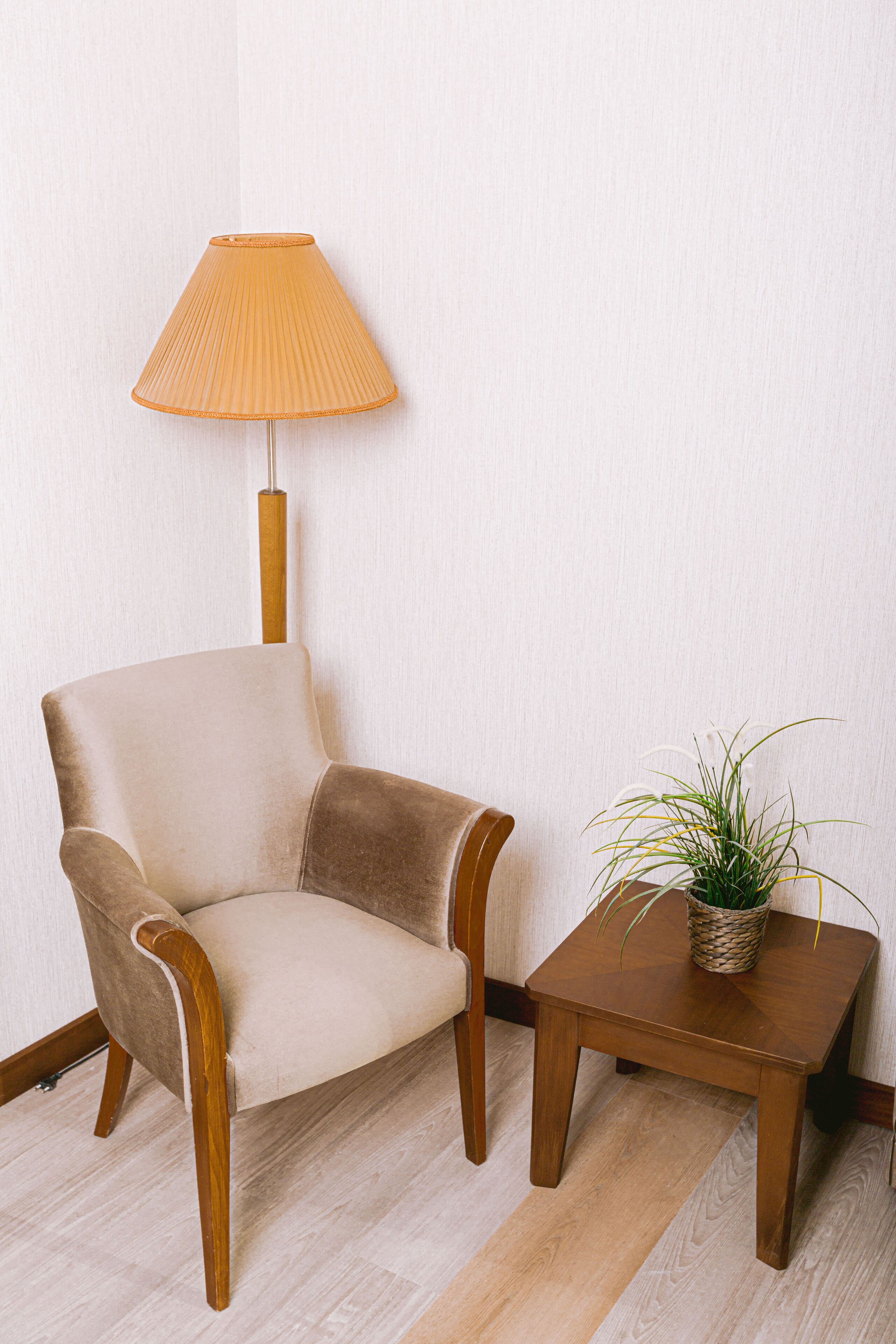 Ett rum med en beige fåtölj, ett litet träbord med en växt och en lampa på ett trägolv.