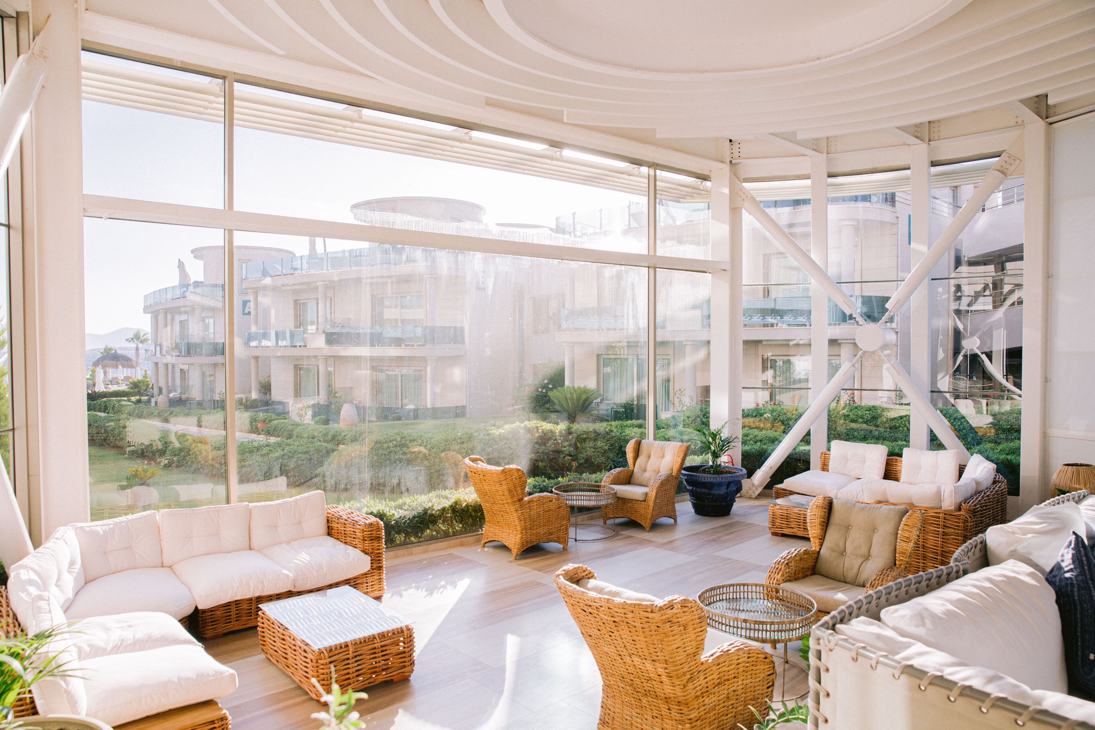 A spacious, well-lit room with large windows offering a view of a modern building and greenery. There are multiple wicker sofas and chairs arranged for seating.