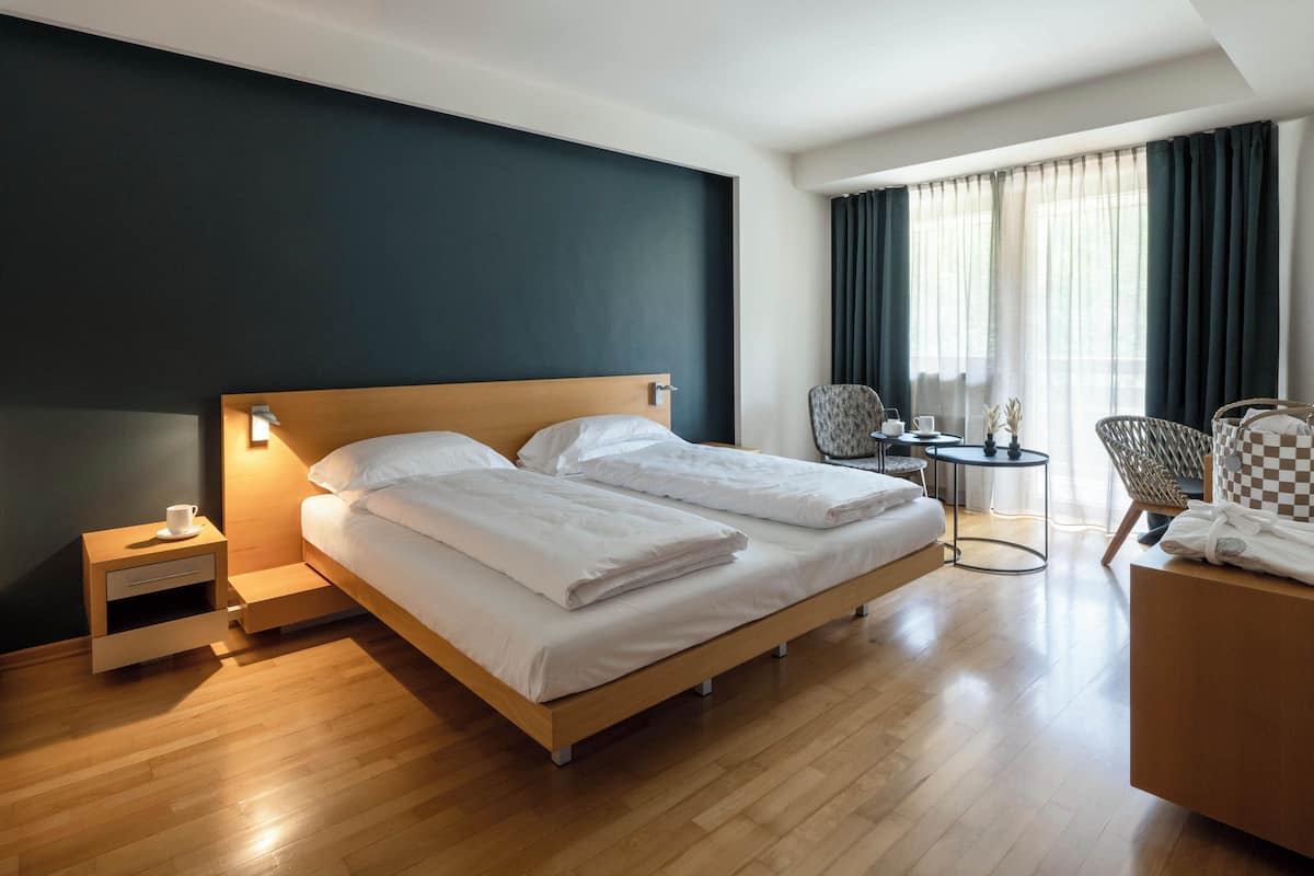 A hotel room with a bed, bedside table, chair, and a small table with a lamp.