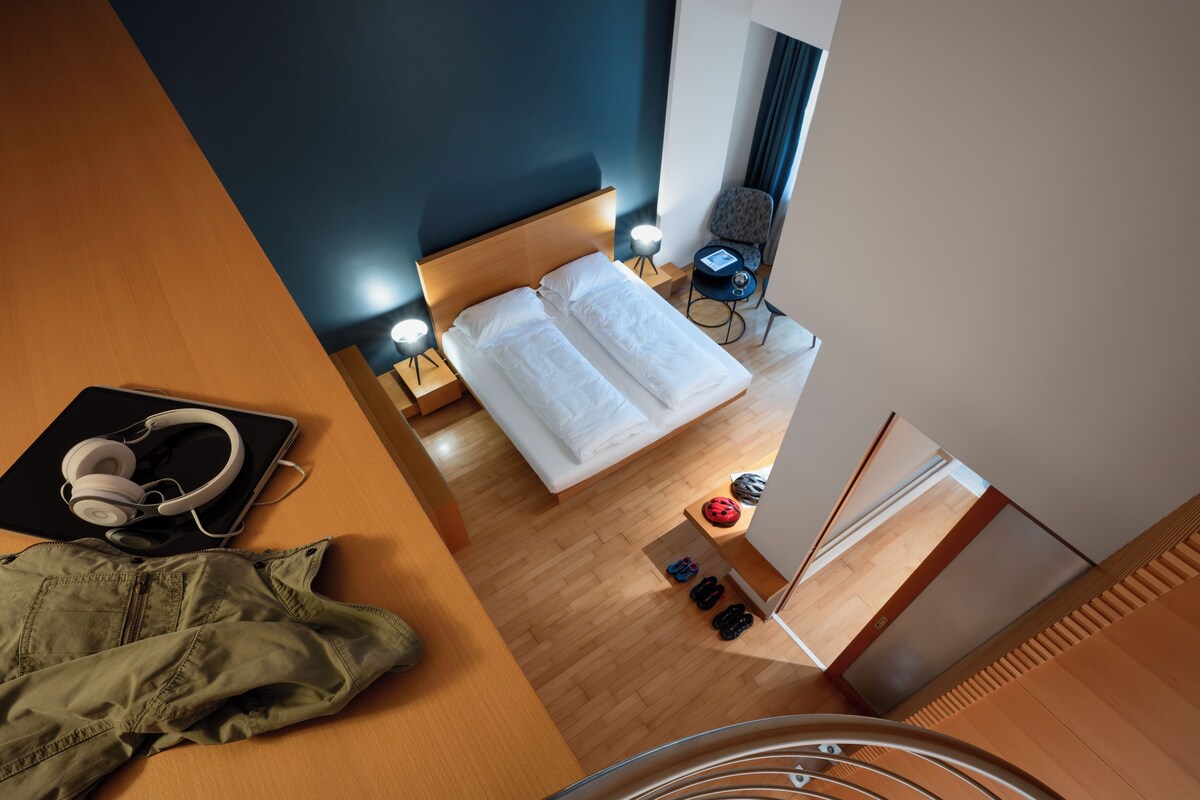 A hotel room with a bed, bedside tables, a chair, and a desk with headphones and a jacket.