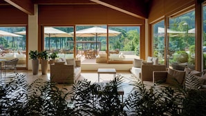 A spacious indoor area with large windows, wicker furniture, and potted plants. - Grand Hotel Terme di Comano (Stenico)