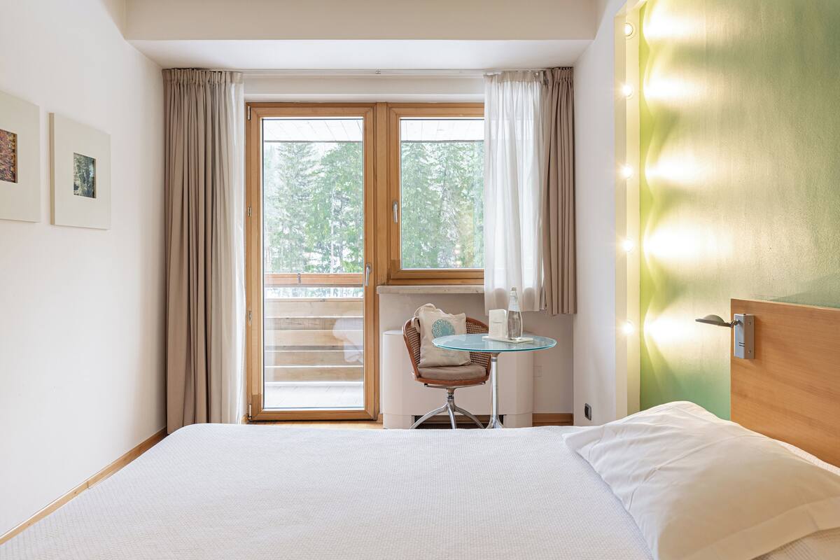 A hotel room with a bed, a small table with a chair, a window with curtains, and a view of trees outside.