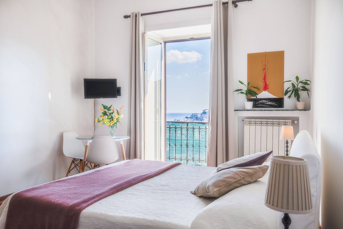 Ein Hotelzimmer mit Bett, Schreibtisch, Fernseher, einem Balkon mit Meerblick und einem Bild an der Wand.
