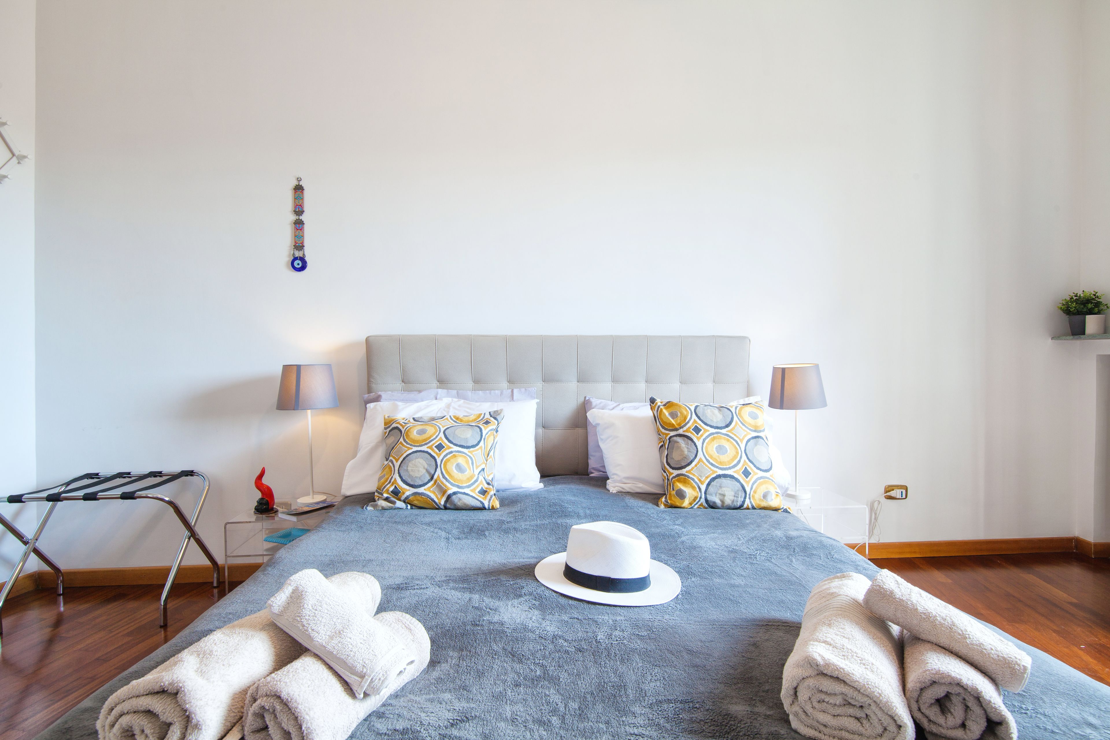 A neatly made bed with a grey blanket, two patterned pillows, a white hat, and rolled towels on a wooden floor.