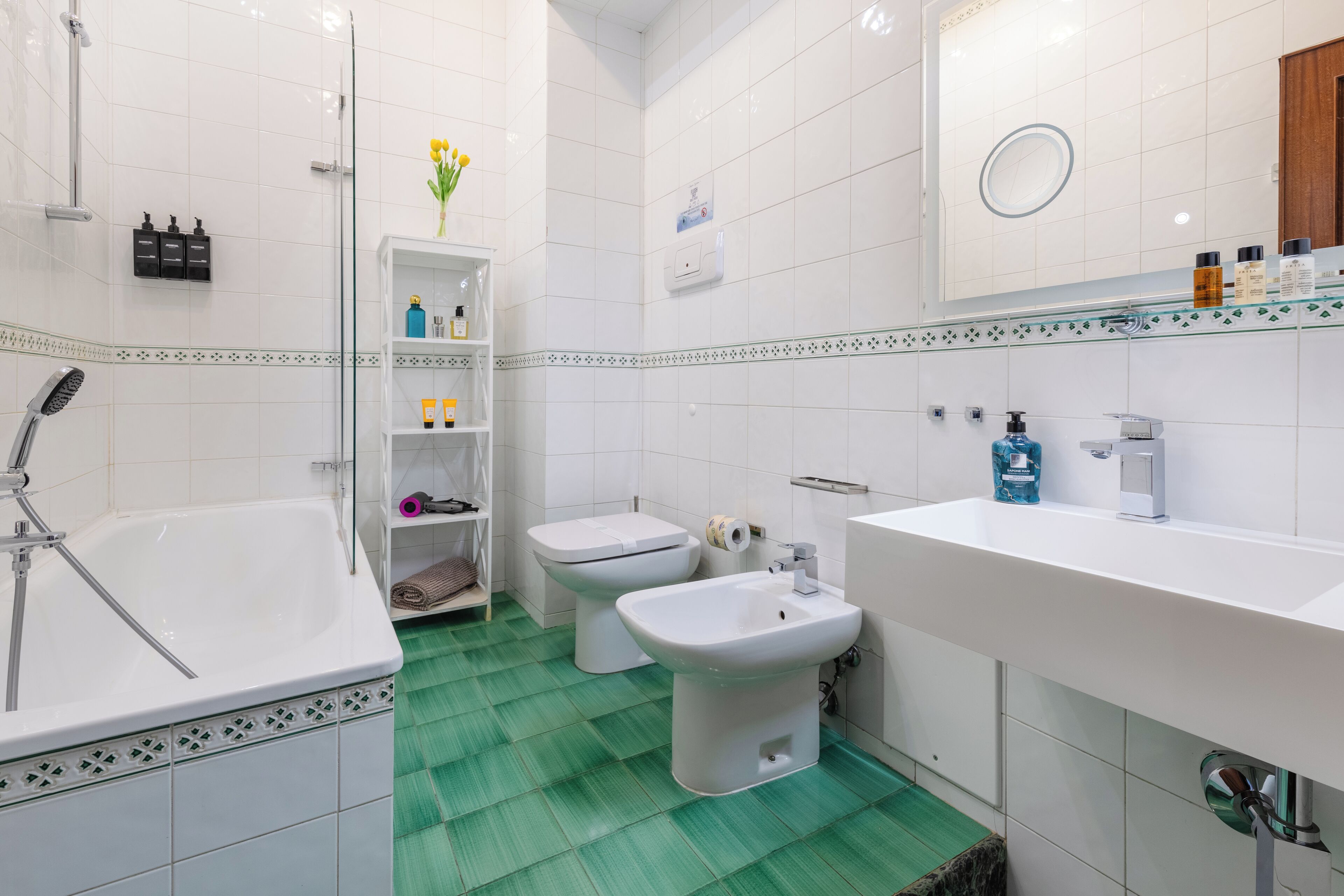 A bathroom with a bathtub, bidet, and sink, with toiletries on shelves and a mirror above the sink.