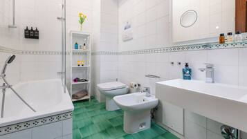 A bathroom with a bathtub, bidet, and sink, with toiletries on shelves and a mirror above the sink.