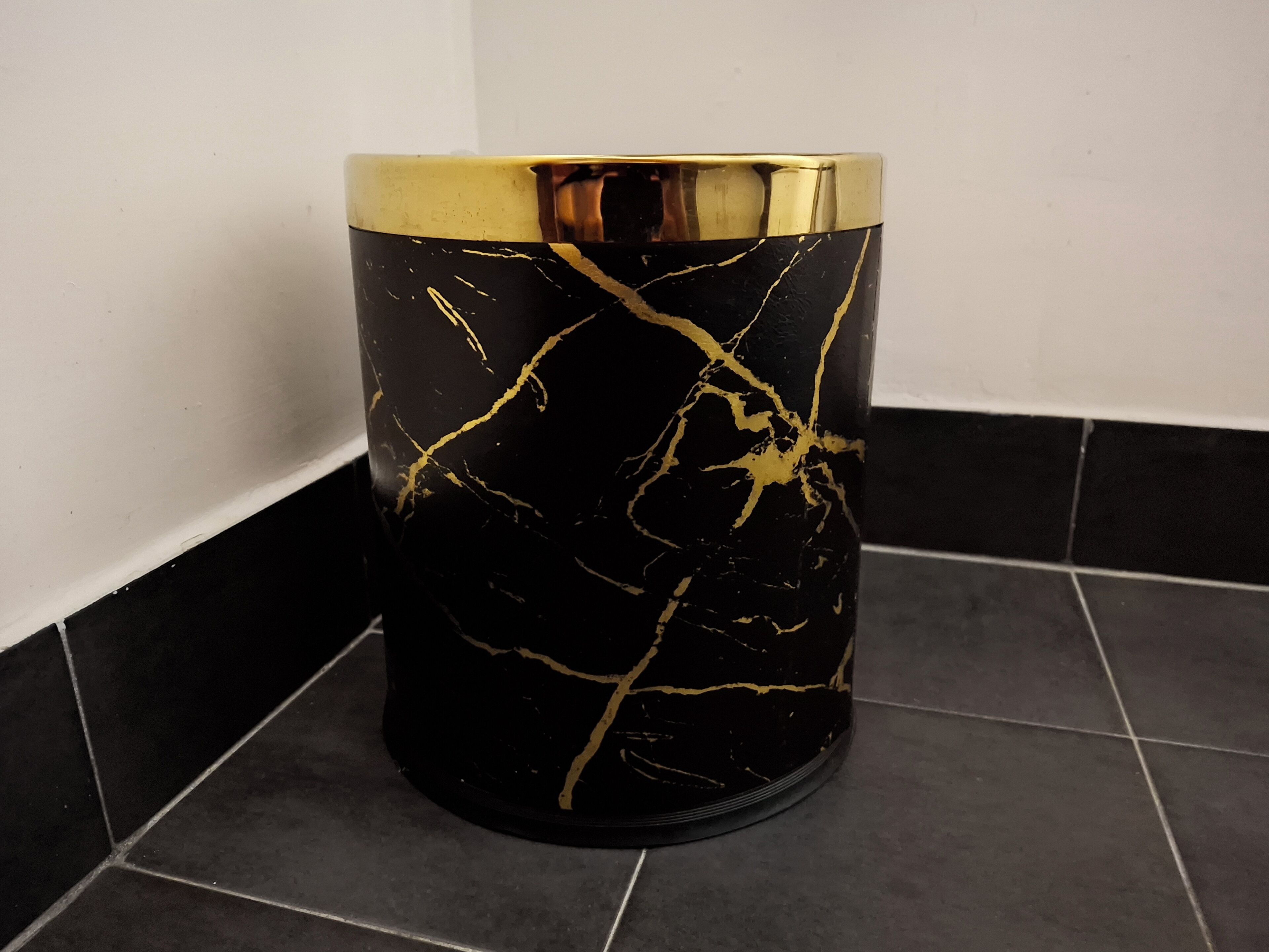 Une poubelle cylindrique à la base noire et aux détails dorés, posée sur un sol carrelé.