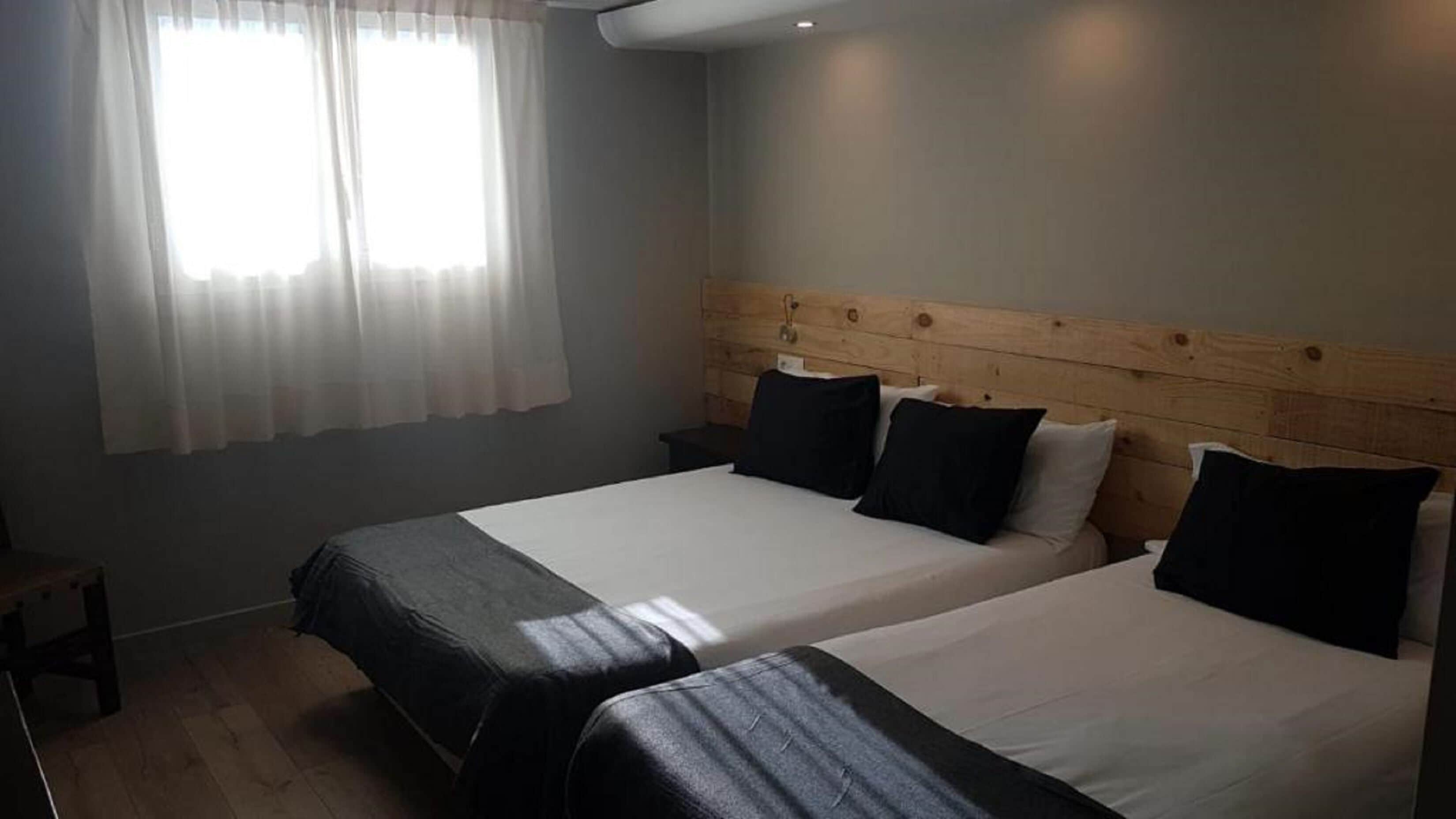 Una habitación con dos camas, cabecero de madera, mesita de noche y ventana con cortinas translúcidas.