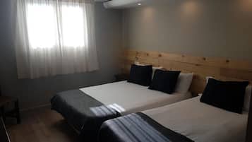Una habitación con dos camas, cabecero de madera, mesita de noche y ventana con cortinas translúcidas.