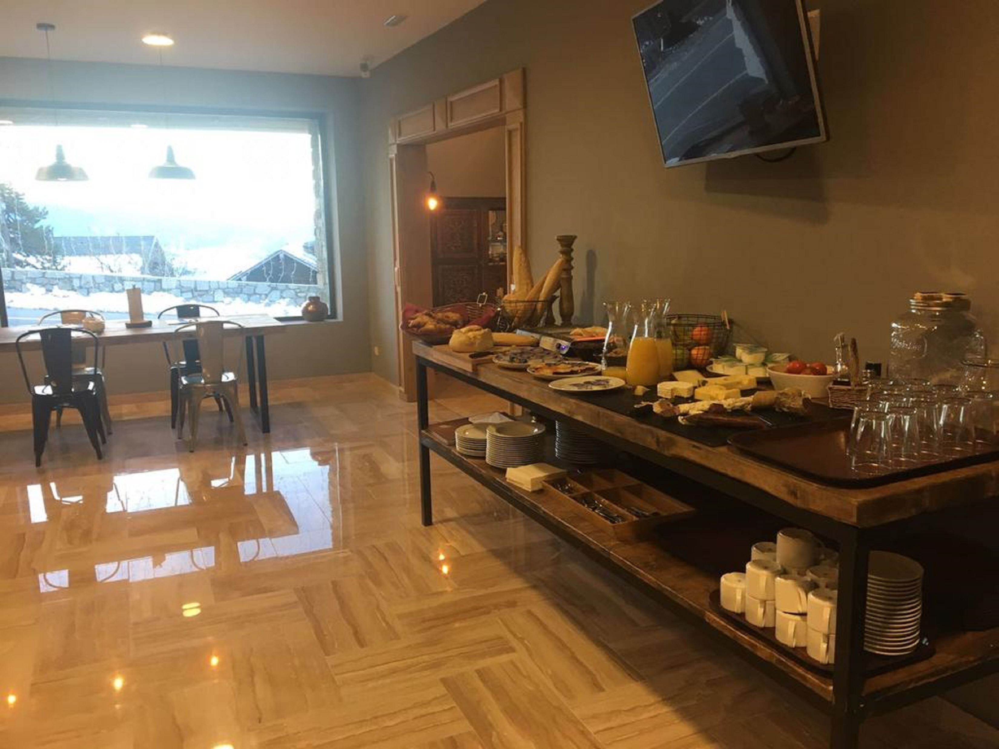 Un buffet de petit-déjeuner avec divers plats, assiettes et verres disposés sur une table en bois, avec des chaises et un téléviseur accroché au mur.
