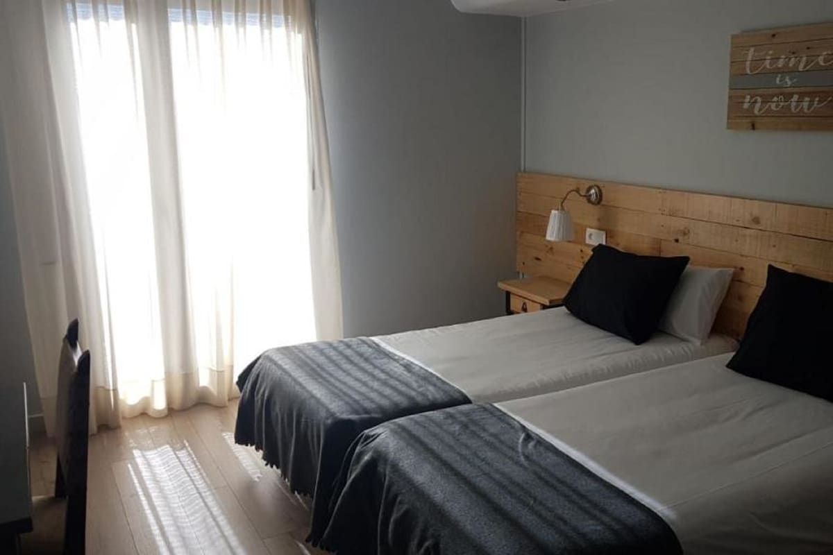 A double room with two beds, a wooden headboard, a bedside table with a lamp, and a window with curtains.