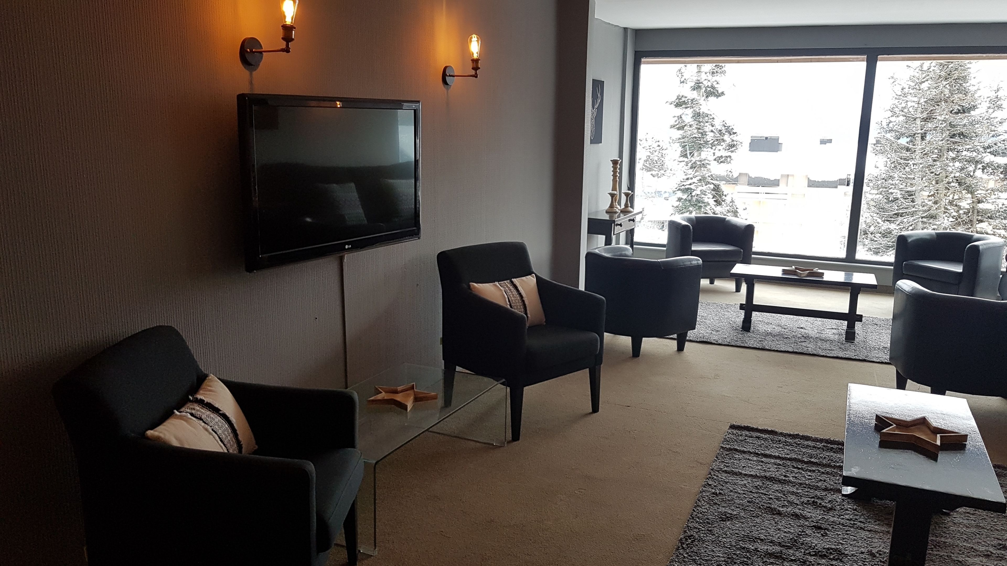 Una sala de estar moderna con una televisión de pantalla plana, sillas tapizadas en negro, una mesa de centro de vidrio y un ventanal con vistas a árboles y edificios.