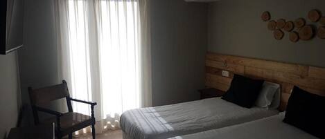 A hotel room with a bed, a chair, a bedside table, a wall-mounted clock, and a window with sheer curtains.