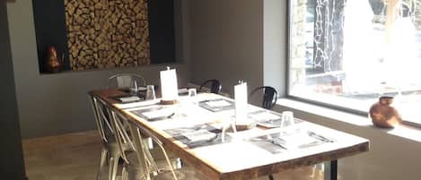 A dining area with a long wooden table set for six, surrounded by metal chairs. There are pendant lights above the table, and a stone wall with a fire feature to the left.