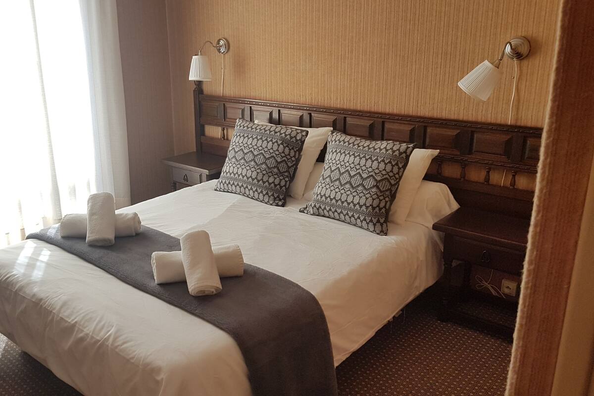 Habitación de hotel con una cama grande, dos lámparas de noche, un cabecero de madera y una ventana con cortinas.