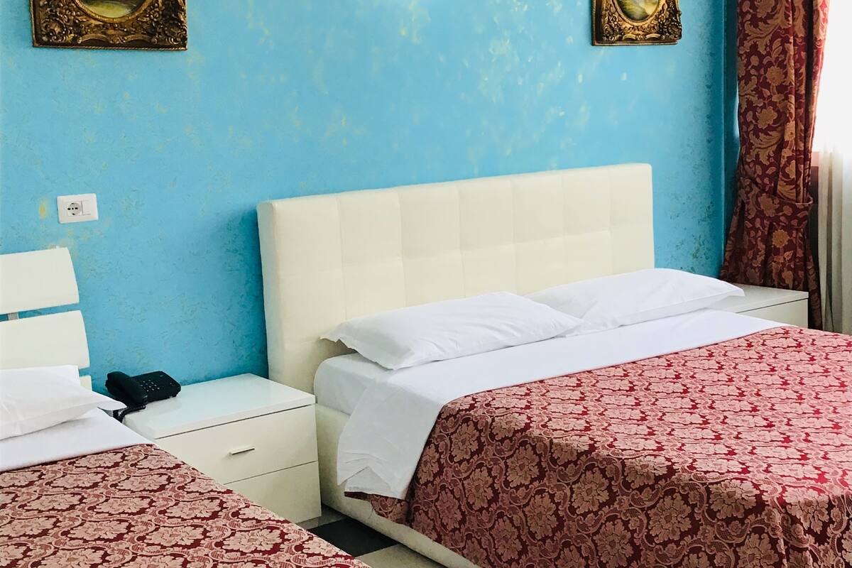 Una habitación de hotel con dos camas, cabecero blanco, colchas estampadas, dos cuadros en las paredes, una mesita de noche blanca con un teléfono y suelo a cuadros.