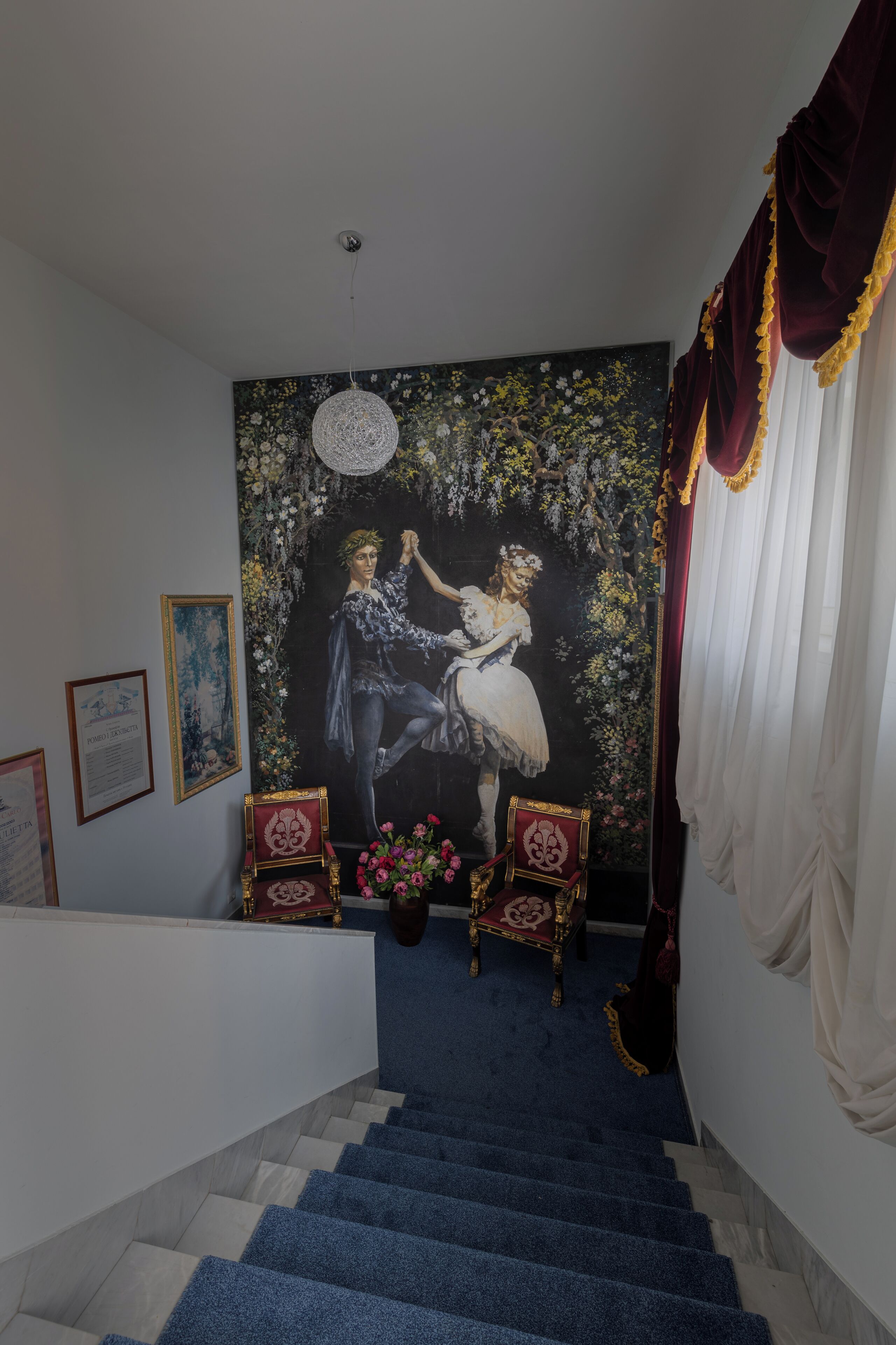 A staircase with blue carpet leading to a room with a large floral tapestry, two ornate chairs, and a chandelier.