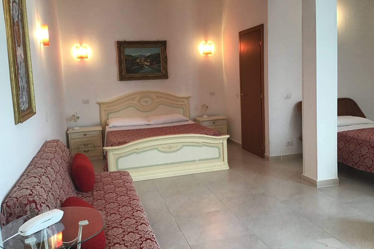 Un dormitorio con cama, mesita de noche, lámpara, sofá y un cuadro en la pared.