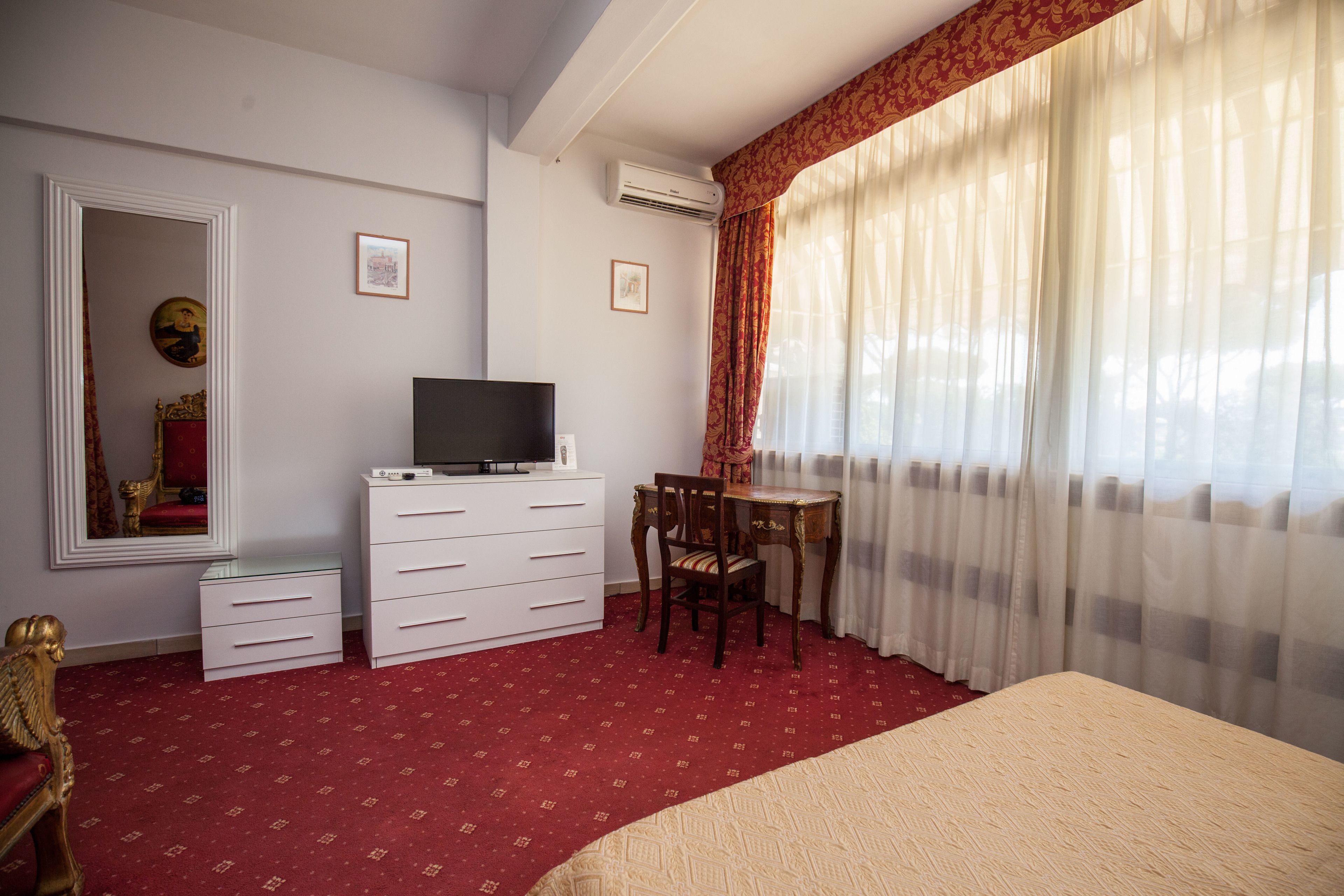 A hotel room with a bed, a TV on a dresser, a desk with a chair, and a large window with sheer curtains.