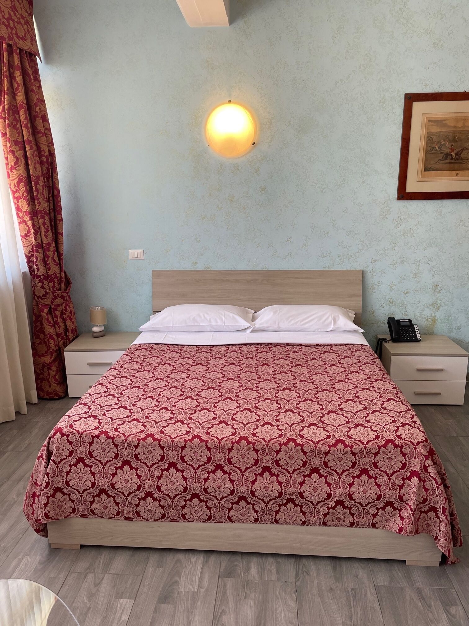 A neatly made bed with a patterned bedspread, a bedside lamp, a nightstand with a phone, and a wall-mounted light fixture.
