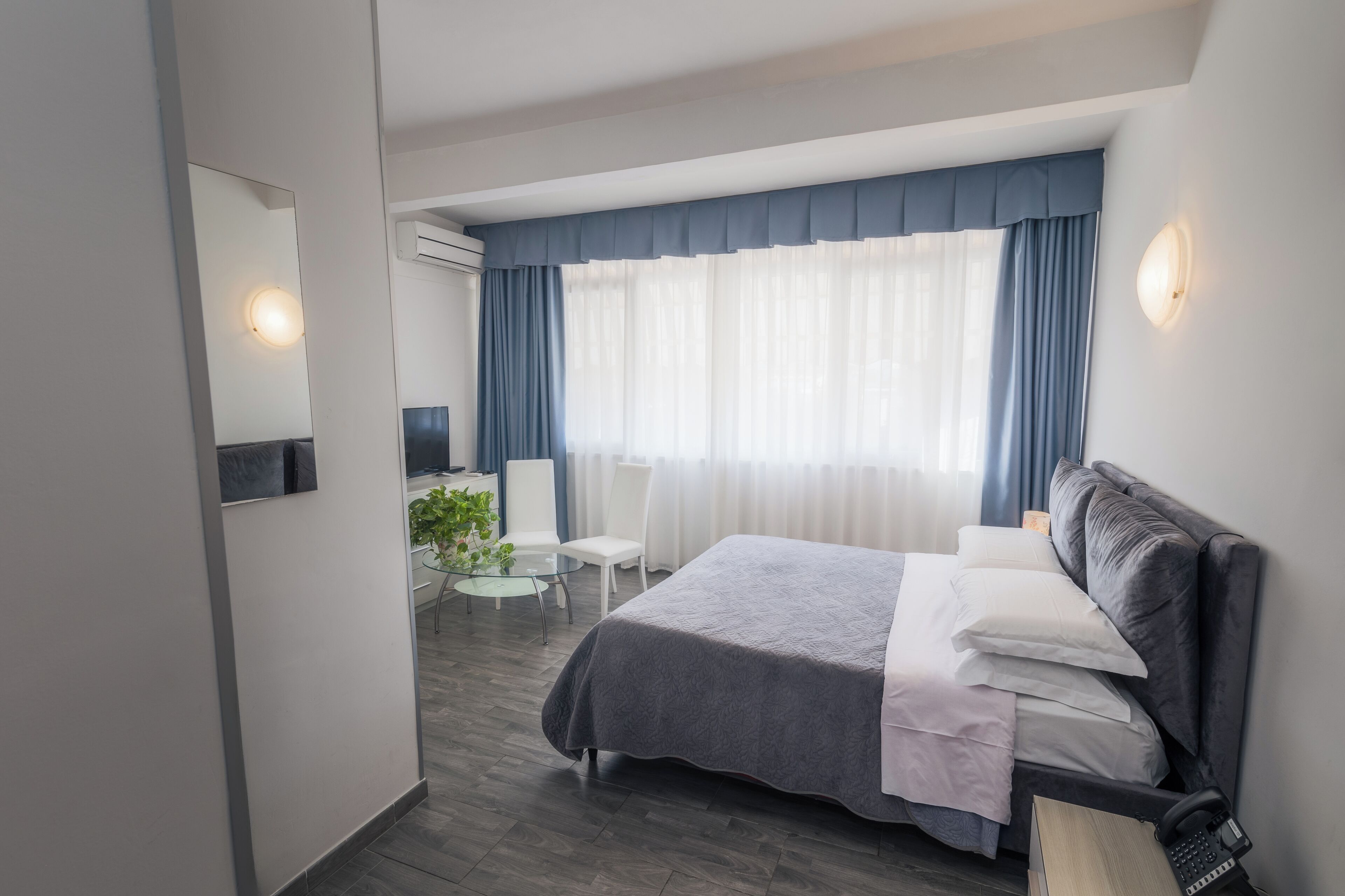 Een moderne hotelkamer met een groot bed, een bureau met stoel, een kleintje tafeltje met een plant en een groot raam met gordijnen.