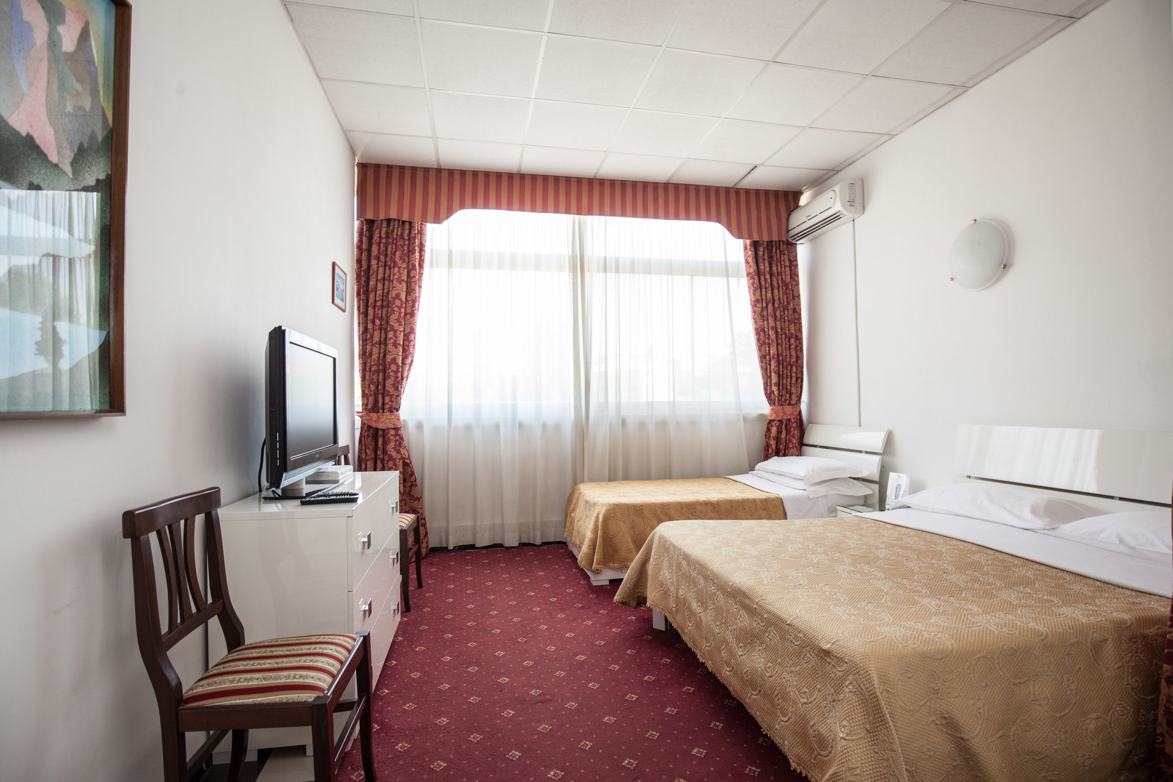 A hotel room with two beds, a desk with a television, a chair, and a large window with curtains.