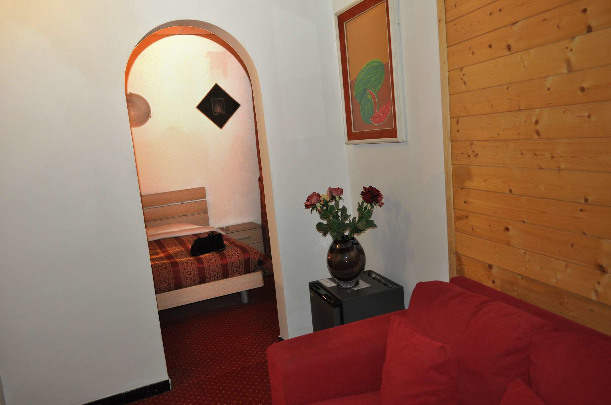 A small room with a bed, a red sofa, and a vase with flowers.