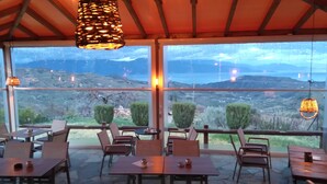 Un área de comedor al aire libre con mesas y sillas, ventanas amplias que ofrecen vista a un paisaje montañoso.