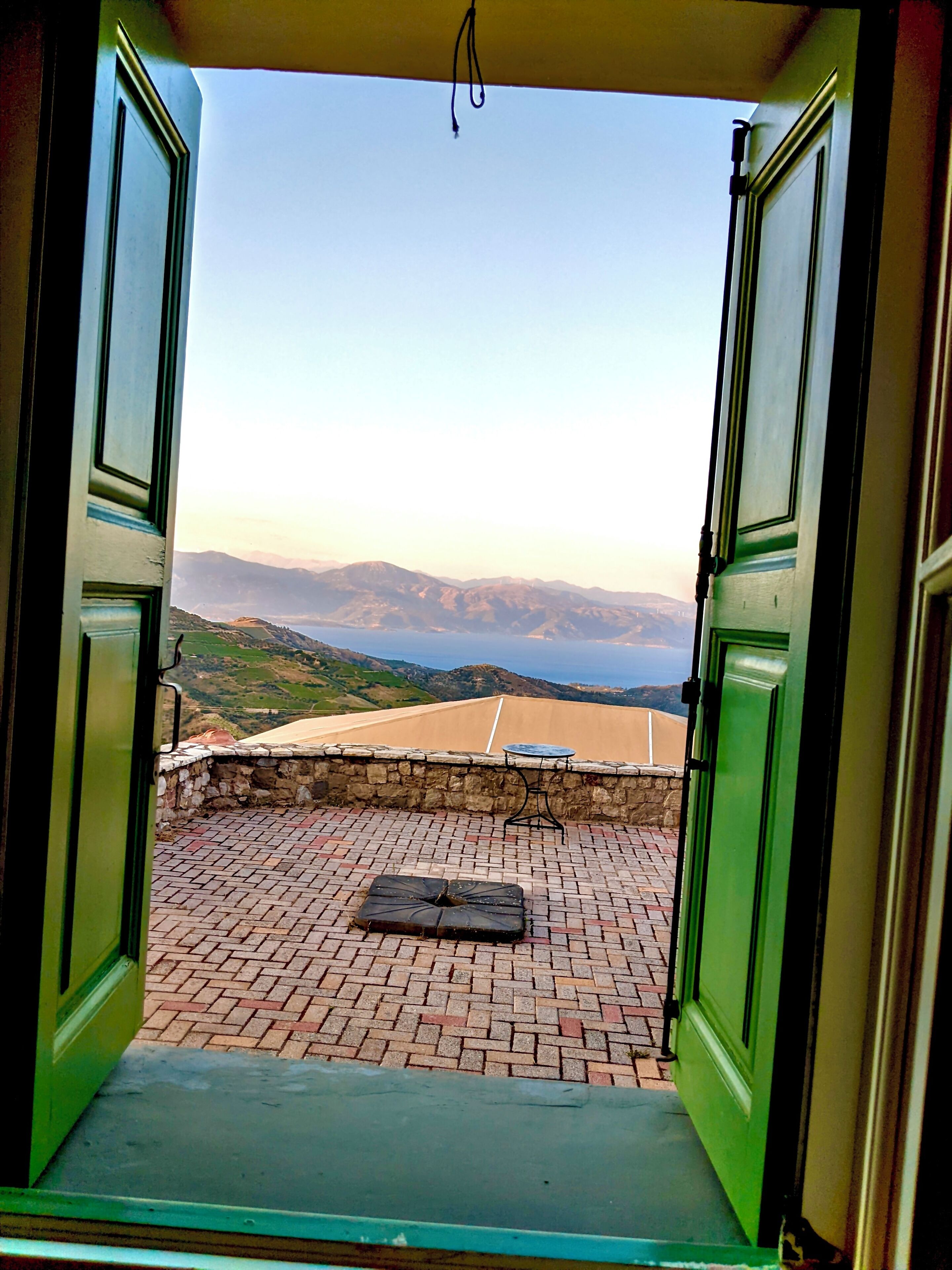 An open door leading to a terrace with a brick floor, a table, and a view of distant mountains and a body of water.
