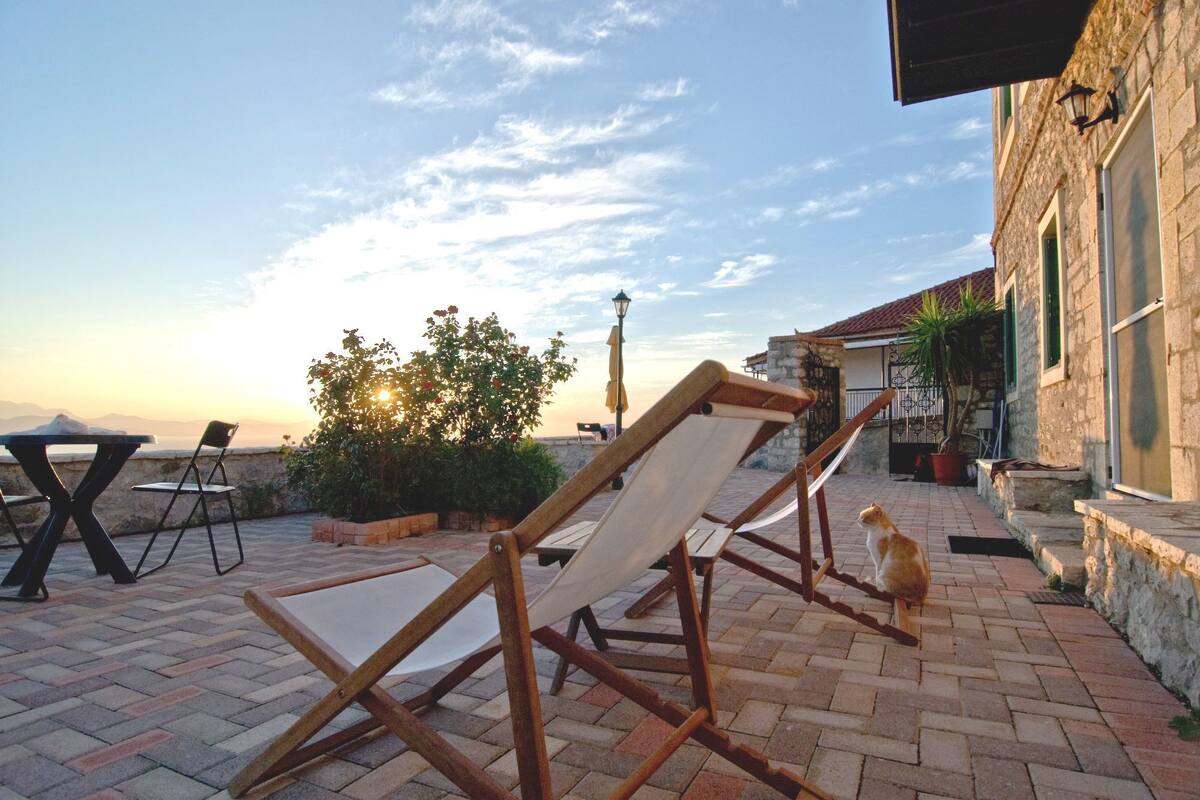 A patio with lounge chairs, a table, and a stone building with a view of the sea.