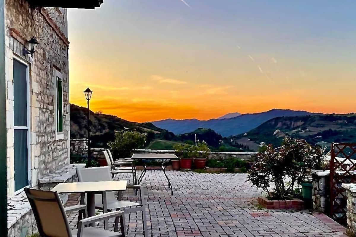 A patio with brick flooring and outdoor furniture, overlooking a scenic landscape with mountains and a sunset.