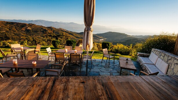 Un área de comedor al aire libre con suelo de piedra, mesas y sillas de madera, con vistas a un paisaje montañoso.