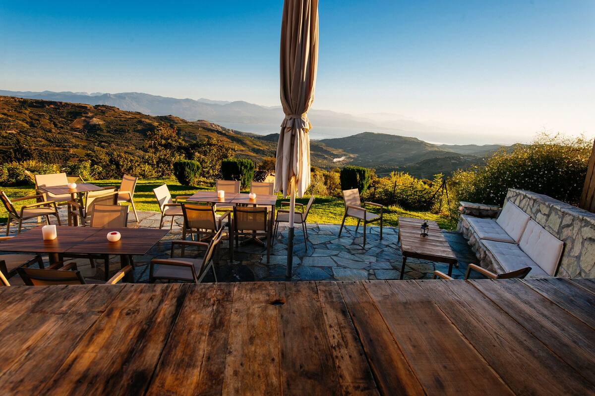An outdoor dining area with stone flooring, wooden tables, and chairs, overlooking a mountainous landscape.