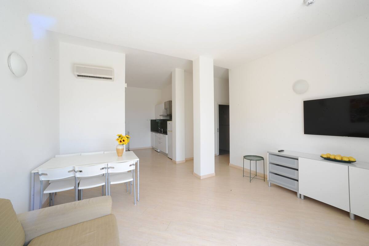 Una sala de estar moderna y minimalista con una mesa de comedor blanca, un sofá, un televisor de pantalla plana y una cocina al fondo.