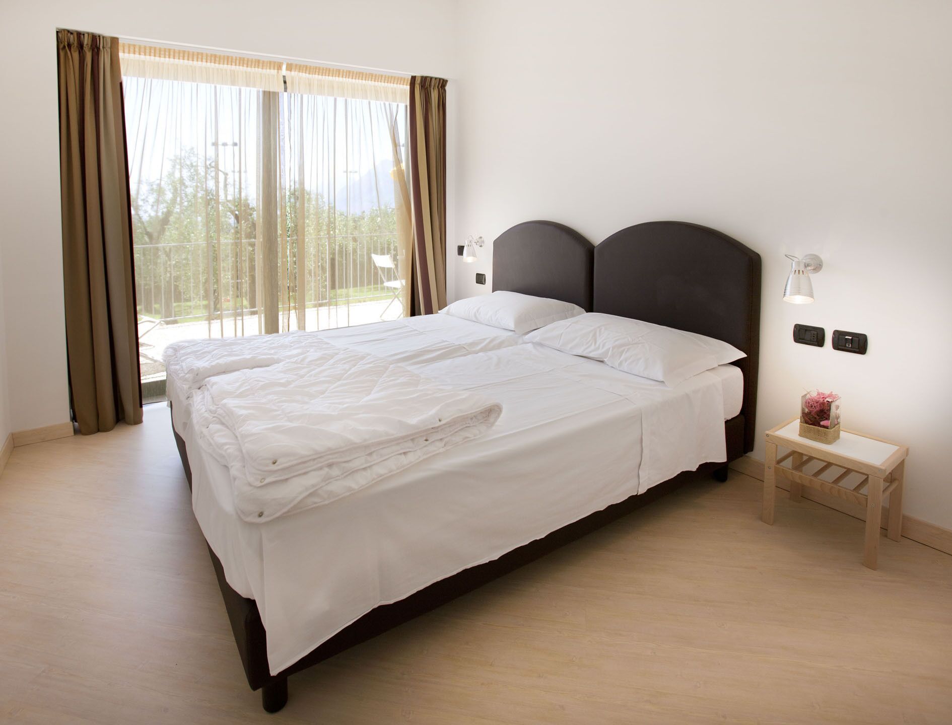 Una cama doble con cabecera oscura en una habitación con un ventanal grande, una mesita de noche pequeña y un aplique de pared.